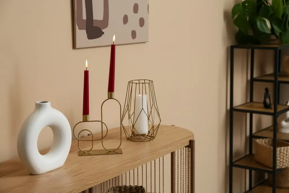 Une étagère minimaliste en bois supporte un vase blanc circulaire, deux bougies rouges allumées dans un bougeoir moderne, une lanterne géométrique en fil de fer et un tableau au-dessus.