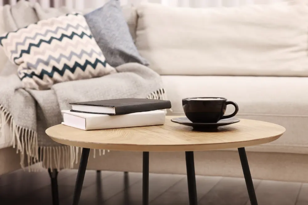 Une scène de salon chaleureuse avec un canapé beige clair, des coussins à motifs en zigzag et un plaid gris. Sur une table en bois reposent deux livres et une tasse à café noire.