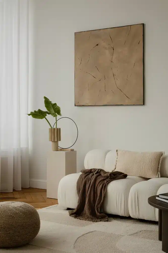 Un salon minimaliste avec un canapé blanc, un plaid beige texturé et un coussin. Au-dessus, une peinture abstraite aux tons neutres. À gauche, une plante dans un vase moderne. Ambiance chaleureuse et sereine.