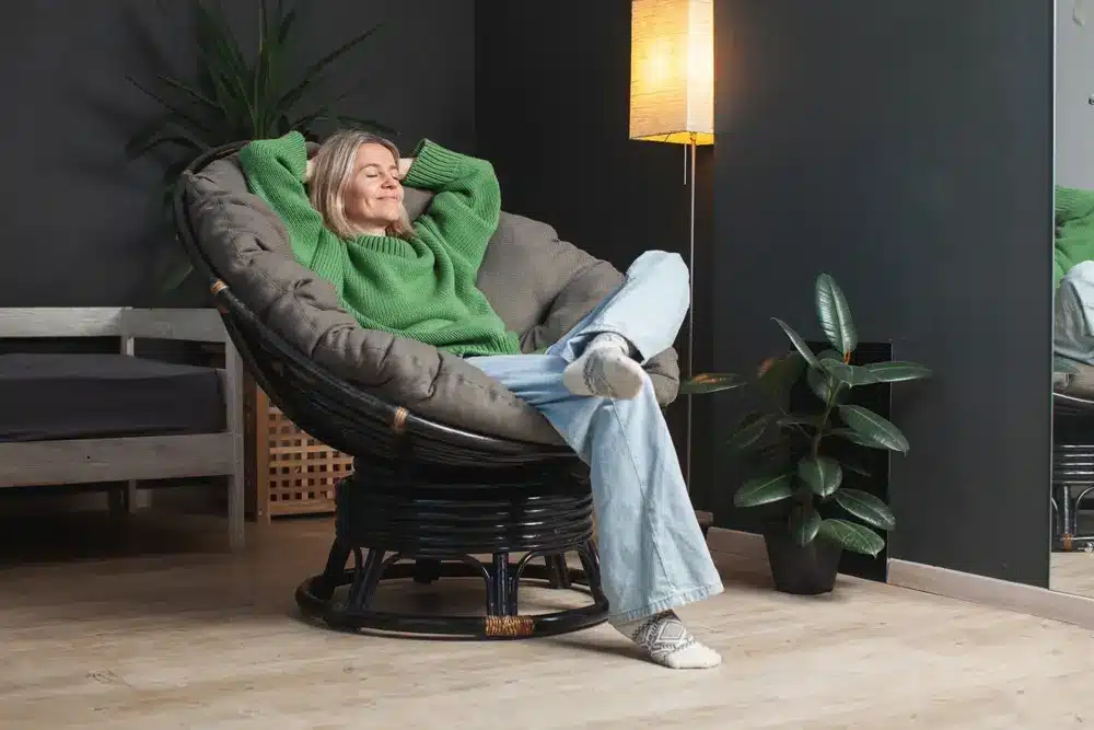Une femme en pull vert et jean se détend dans un fauteuil papasan, souriant les yeux fermés. À proximité, une plante en pot et un lampadaire créent une ambiance chaleureuse.