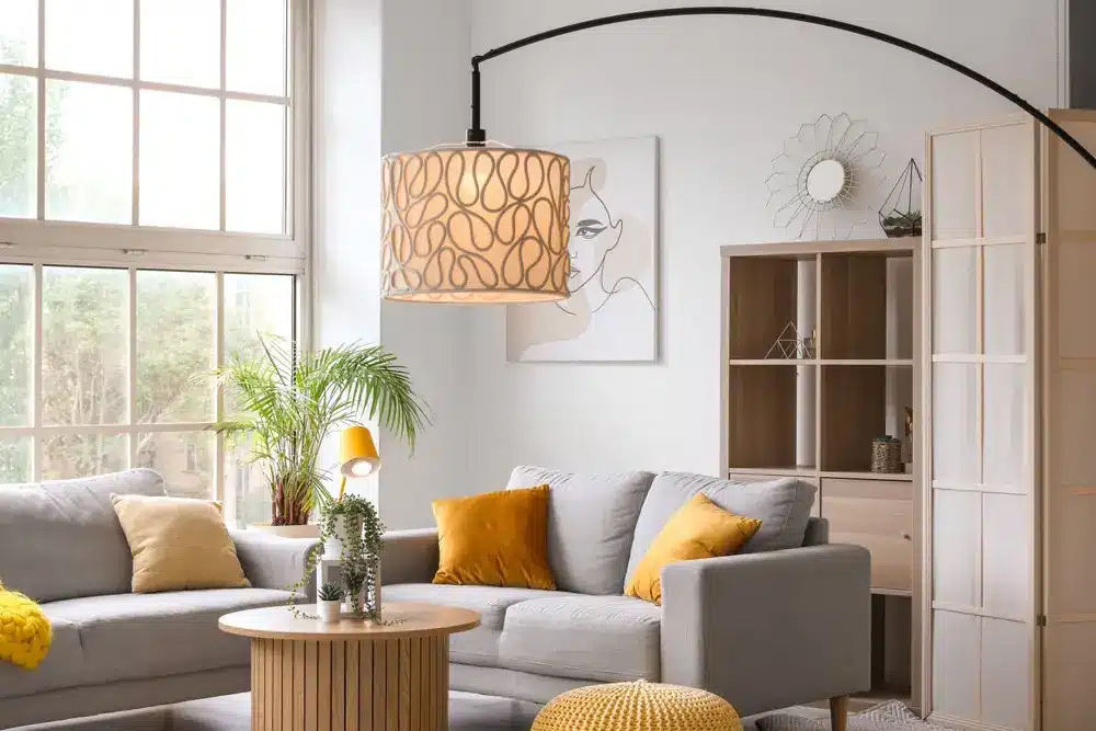 Salon moderne avec un canapé gris, des coussins jaunes et une table ronde. Une grande fenêtre, des œuvres d'art abstraites et un lampadaire incurvé créent une ambiance chaleureuse.