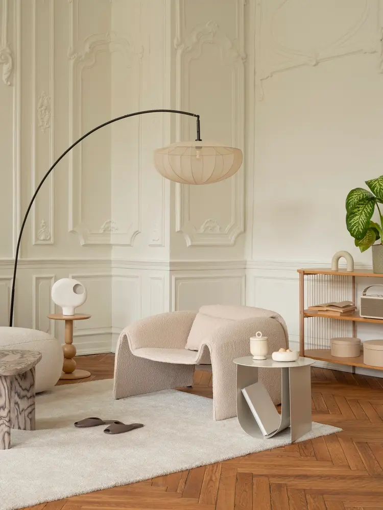 Salon raffiné avec un élégant fauteuil beige, un lampadaire moderne et une table d'appoint épurée sur un tapis moelleux. Des touches de bois naturel et une plante verte apportent une note chaleureuse.
