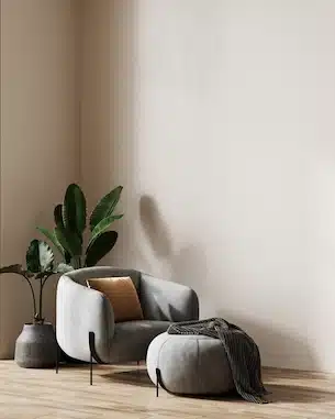 Une chambre minimaliste comprend un fauteuil gris avec un coussin moutarde et un repose-pieds assorti, une couverture drapée et une plante à feuilles dans un vase sur un parquet.