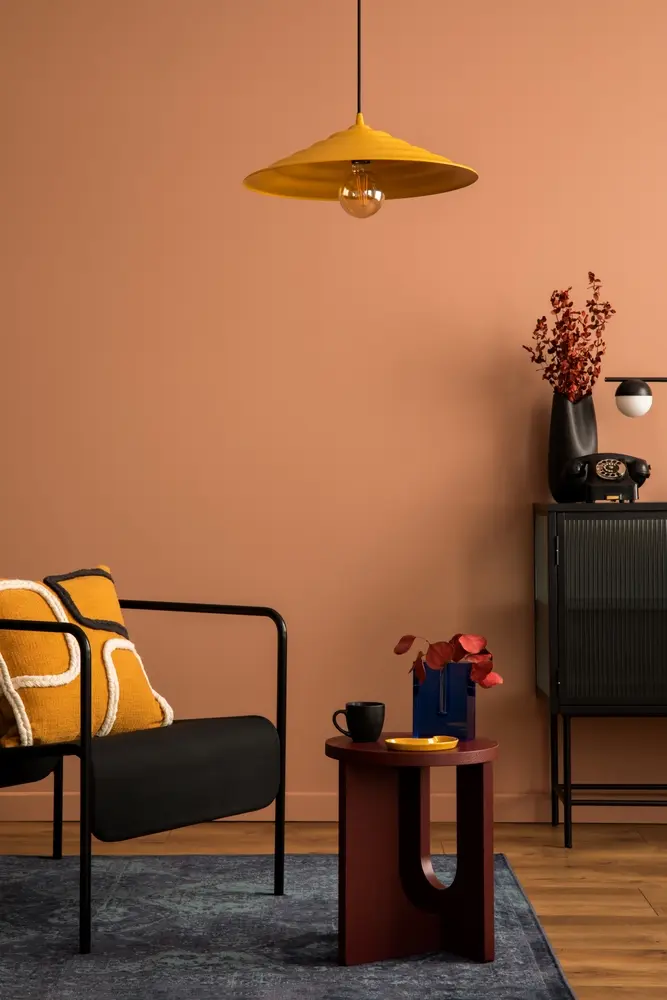 Salon élégant avec un fauteuil noir, des coussins moutarde et une suspension jaune. Sur une table bordeaux, devant un mur brun chaud, se trouvent une tasse noire et un vase de fleurs rouges.