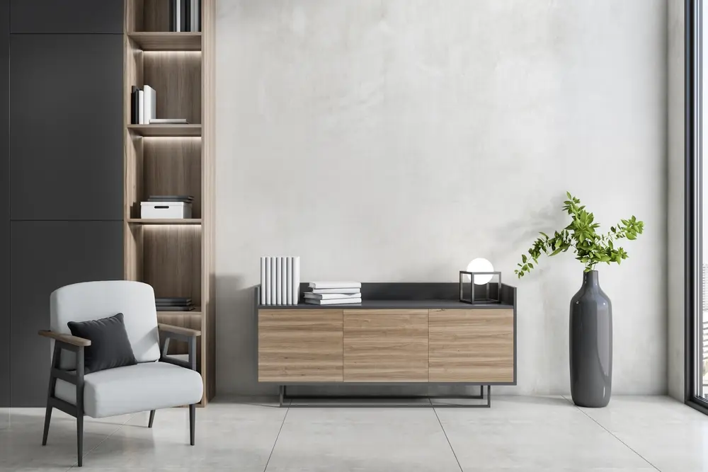 Salon moderne avec un fauteuil confortable, un coussin noir, des étagères en bois élégantes, un buffet avec des livres et un grand vase contenant de la verdure sur un sol en béton.