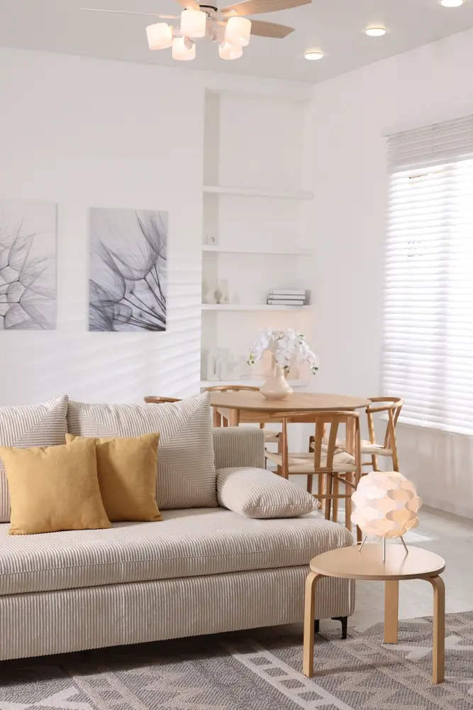 Salon lumineux avec un canapé beige, des coussins moutarde et une table ronde en bois ornée de fleurs. Des œuvres d'art abstraites au mur et un éclairage tamisé créent une ambiance apaisante.