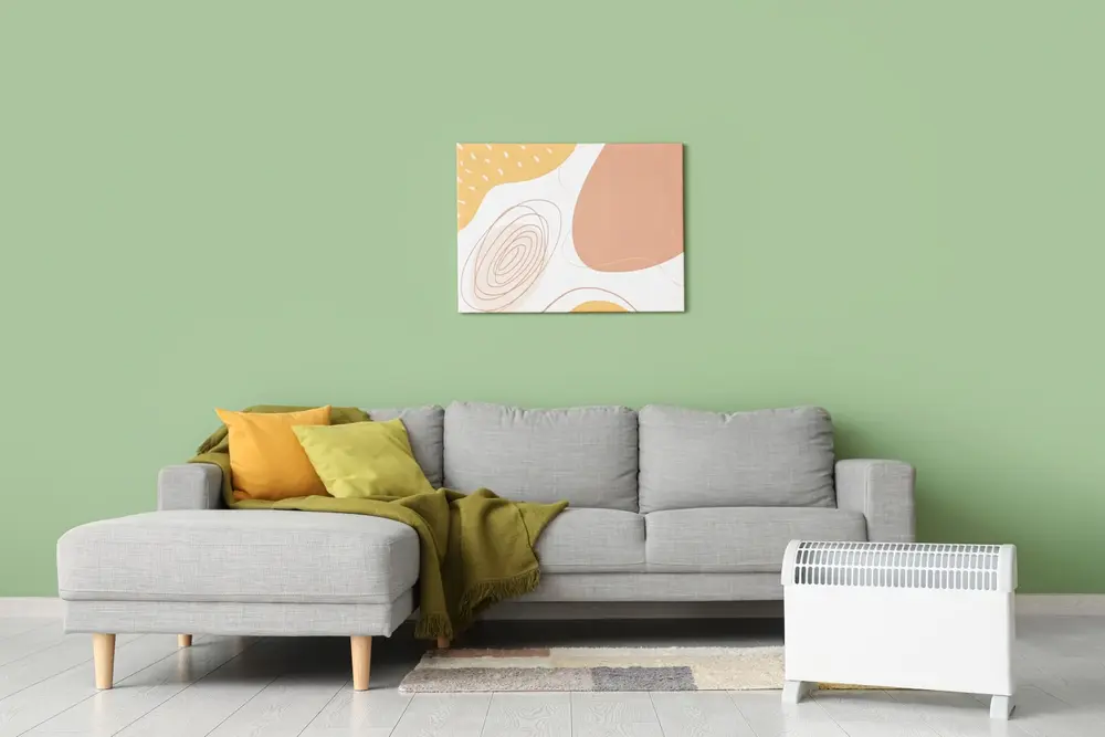 Un salon confortable avec un canapé d'angle gris, des coussins verts et jaunes, un plaid vert, une œuvre d'art abstraite sur un mur vert et un radiateur blanc.