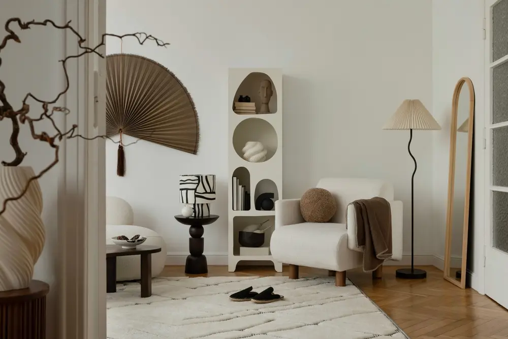 Salon minimaliste aux tons neutres, avec un fauteuil blanc, des étagères rondes, un ventilateur décoratif mural, un lampadaire et des œuvres d'art abstraites. Ambiance chaleureuse et sereine.