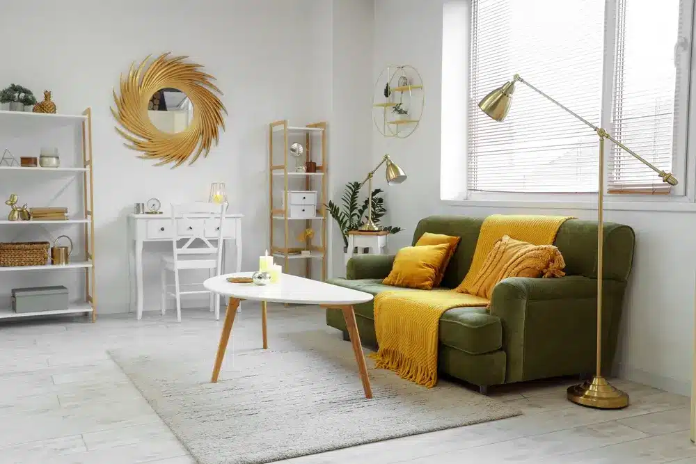 Salon moderne avec un canapé vert, des coussins jaunes et un plaid assorti. Une table basse blanche repose sur un tapis clair. Des touches dorées apportent une note chaleureuse.