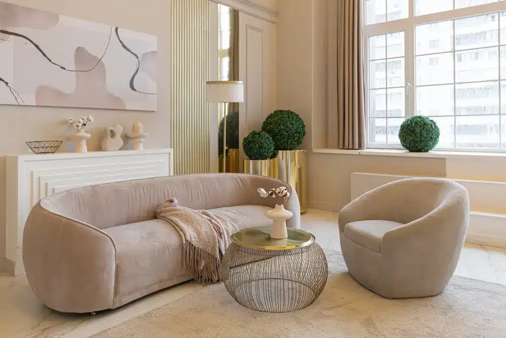 Un salon cosy avec un canapé beige incurvé et un fauteuil assorti, une table basse en fil de fer, des œuvres d'art abstraites au mur, une grande fenêtre et des plantes en pot.
