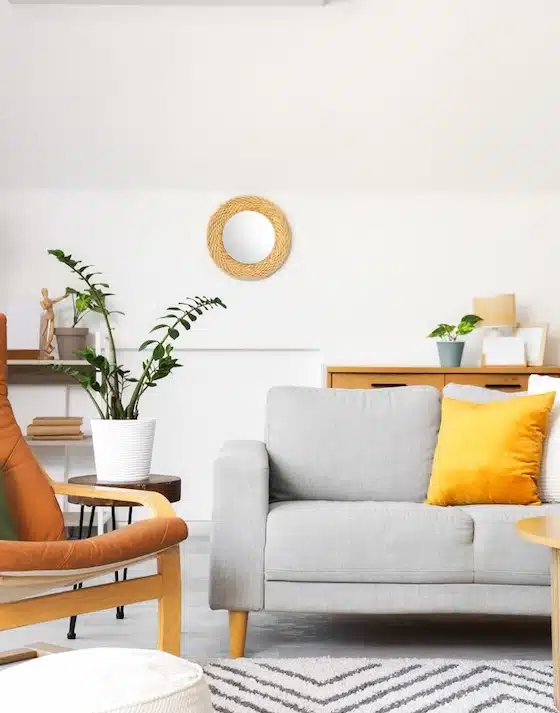 Un salon chaleureux avec un canapé gris orné d'un coussin jaune vif. Un fauteuil en bois au coussin orange se trouve à proximité. Des plantes en pots blancs apportent une touche de verdure, et un petit miroir rond est accroché au mur. L'espace est lumineux et accueillant.