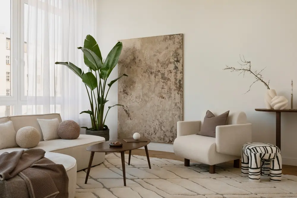Salon moderne avec un canapé crème, des coussins texturés et une grande plante en pot. Art abstrait, décoration minimaliste et lumière naturelle douce créent une atmosphère apaisante.