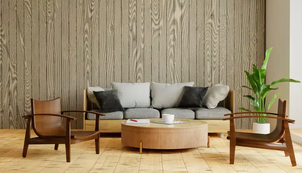 Salon moderne avec un canapé gris et quatre coussins contre un mur lambrissé. Deux fauteuils en bois et une table basse ronde apportent une touche chaleureuse. Une plante en pot ajoute une note de fraîcheur.
