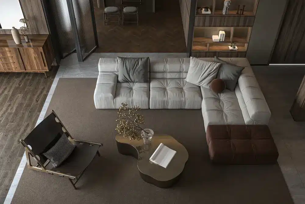Salon moderne avec un canapé d'angle gris, un pouf marron et une table basse sculpturale. Un fauteuil lounge est placé à proximité sur un tapis texturé, créant une ambiance chaleureuse.