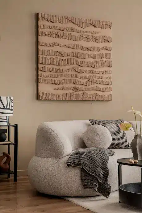 Un salon chaleureux est agrémenté d'un canapé beige arrondi recouvert d'un plaid gris texturé. Au mur, une grande œuvre d'art aux tons naturels, évoquant des vagues, s'harmonise avec le parquet et la décoration épurée.