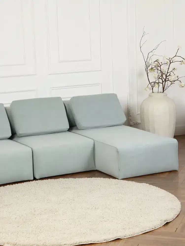 Un canapé modulable bleu clair est adossé à un mur lambrissé blanc, à côté d'un grand vase aux branches dénudées. Un tapis rond blanc est disposé devant, sur un parquet.