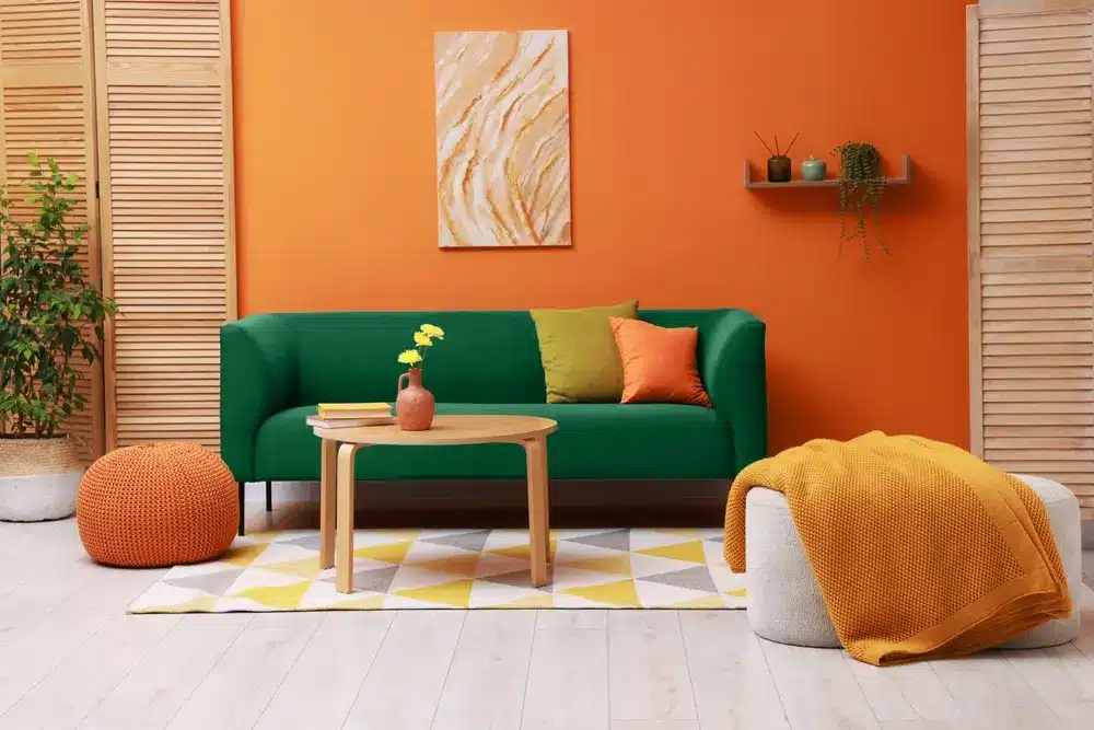 Un salon dynamique avec un mur orange et un canapé vert orné de coussins colorés. Une table basse en bois et des poufs apportent une touche chaleureuse. Des éléments de décoration modernes complètent cette ambiance cosy et accueillante.