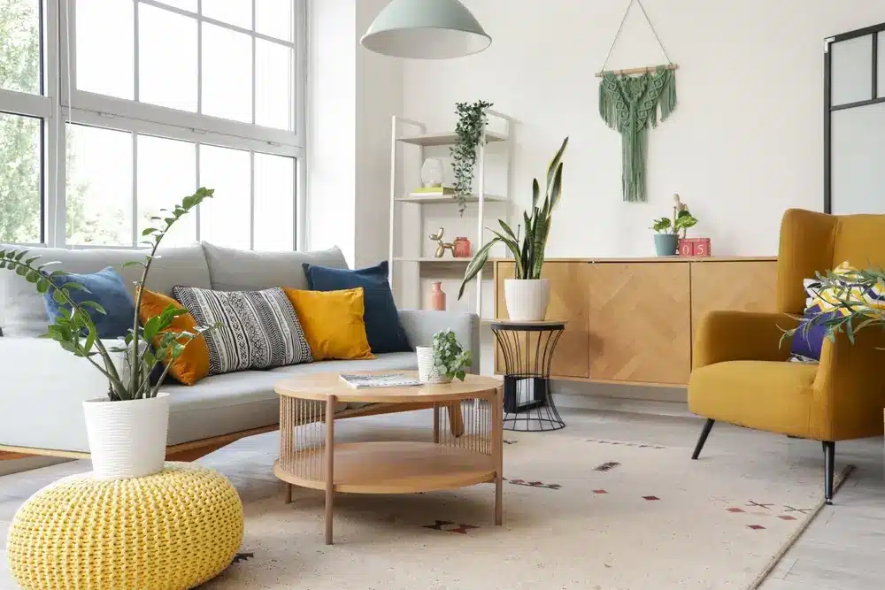 Salon lumineux de style moderne avec un canapé gris clair agrémenté de coussins colorés, un fauteuil moutarde, une table ronde en bois, des plantes et une grande fenêtre.